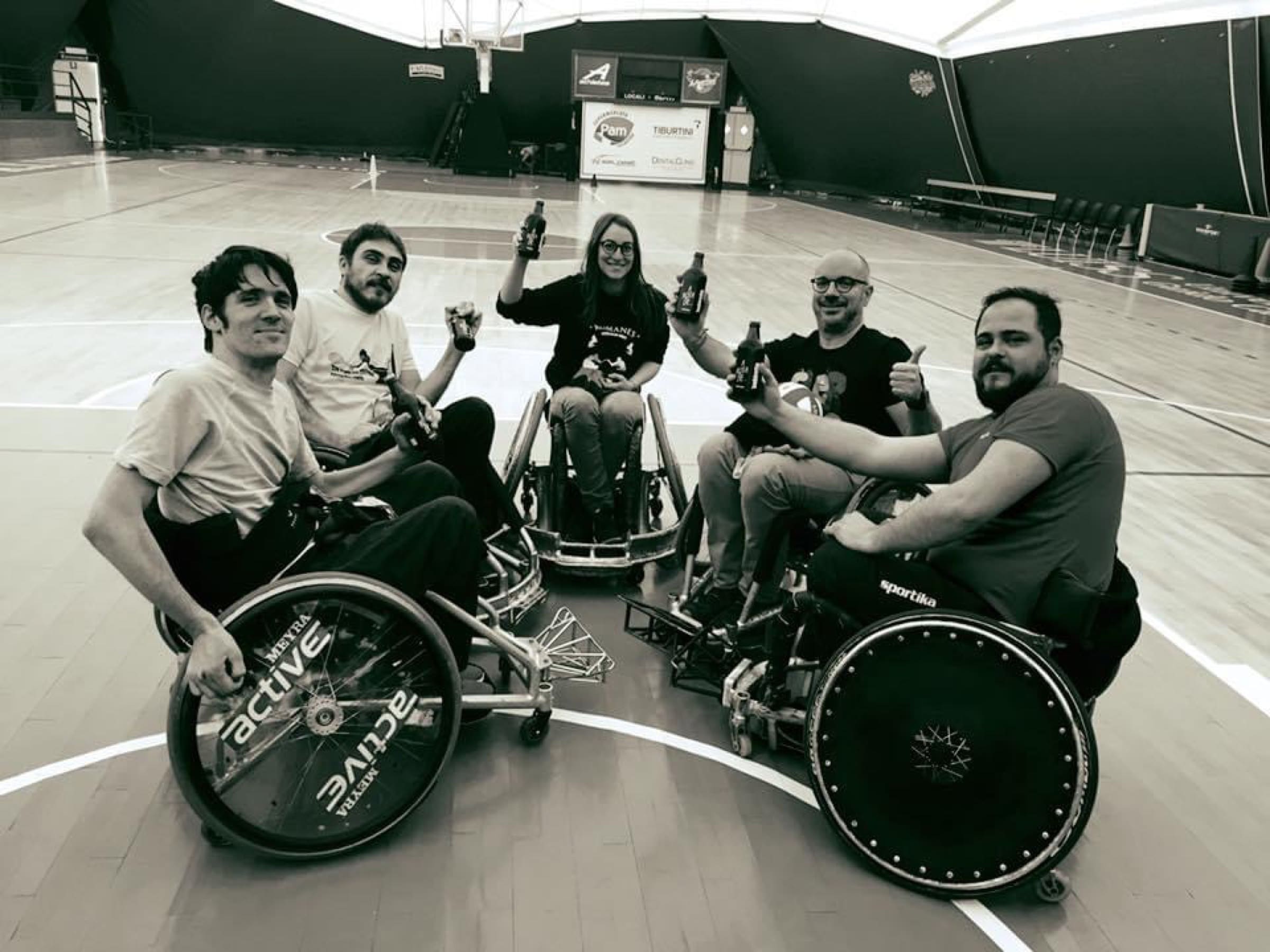Cinque compagni dei Romanes brindano con la birra, in cerchio nelle carrozzine da rugby a centro campo dopo l'allenamento. Foto in bianco e nero.