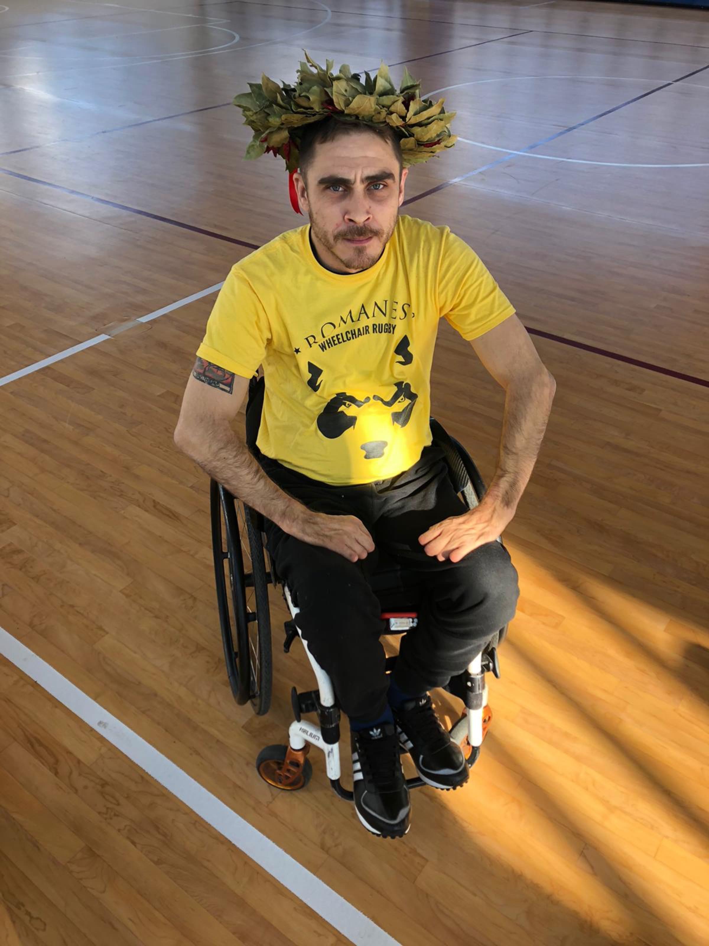 Un giocatore dei Romanes con la maglia gialla e una corona d'alloro in testa, seduto nella carrozzina da rugby in palestra, guarda dritto in camera.