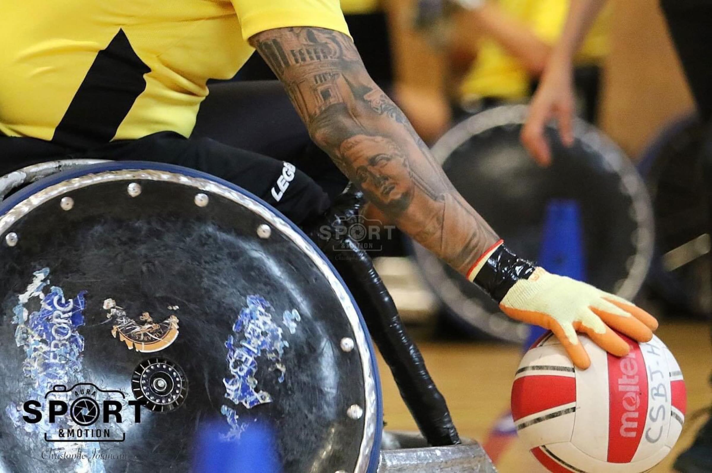 Il braccio tatuato di un giocatore dei Romanes, con il guanto da presa, allunga la mano verso la palla accanto alla ruota corazzata della carrozzina da rugby.